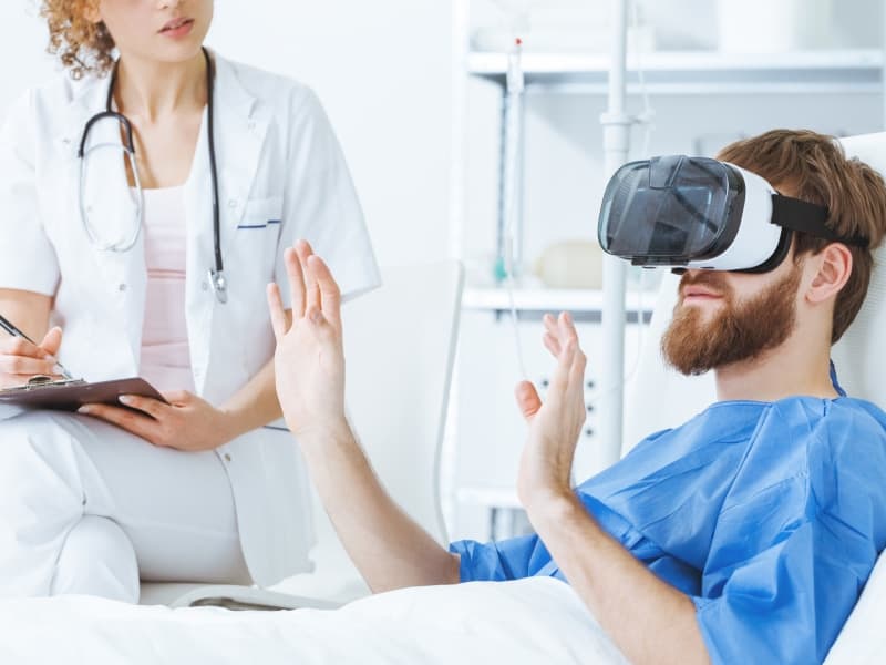 Photograph of a hospital patient in bed wearing virtual reality glasses. A female doctor sits at his bedside taking notes.