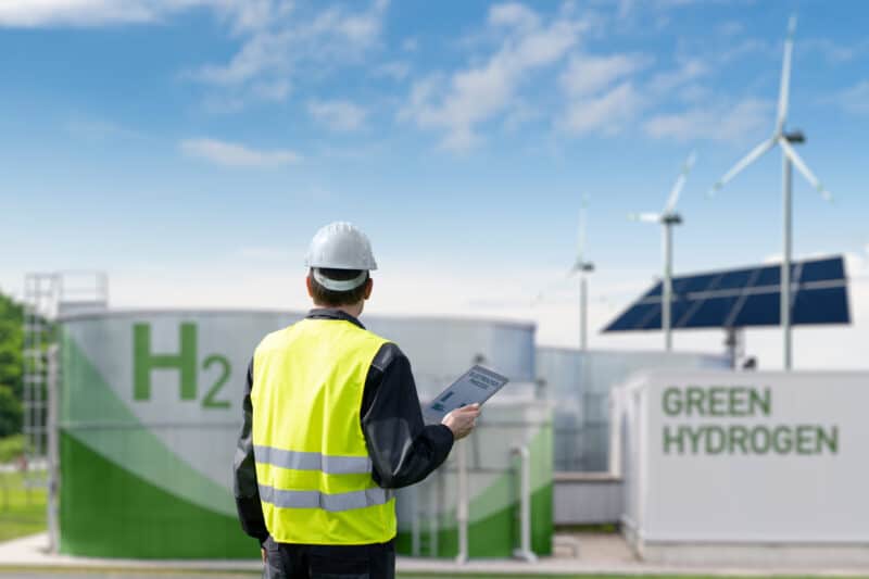 Engineer with a yellow vest and a tablet in his hand stands in front of a green hydrogen factory